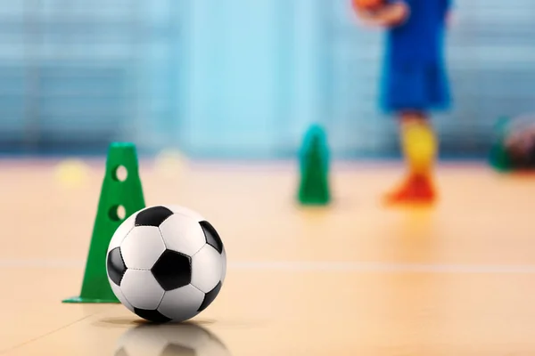 Indoor soccer - futsal training field. Soccer ball and training cones. Kids on practice in blurred background