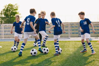 Bir grup çocuk antrenmanda futbol oynuyor. Futbol kulübündeki çocuklar mavi forma ve futbol takımı giyiyorlar. Güneşli bir günde koç ile futbol antrenmanı yapan mutlu çocuklar.