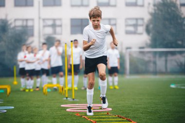 Futbol antrenman kampındaki gençler. Çocuklar çeviklik pratiği yapıyor. Genç sporcular açık hava antrenmanında futbol becerilerini geliştiriyorlar. Oyuncu koşuyor ve merdiven atlıyor. Arka planda okul binası
