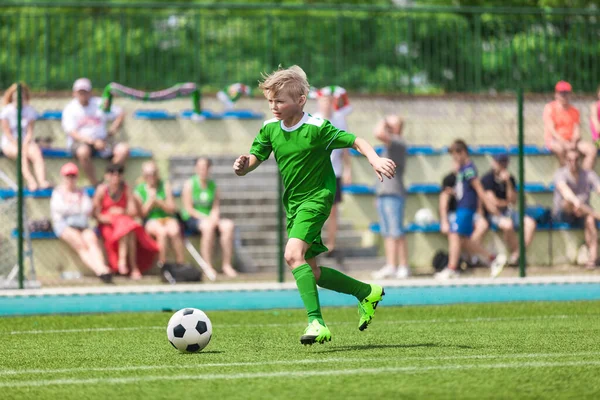School Boy Running Football Ball on Stadium Field. Happy Boy Soccer Player Running Fast Ball on Tournament Competition Match. Kid in Green Sports Jersey Kit: Shirt, Shorts, Socks and Cleats
