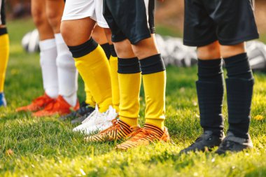Genç futbolculardan oluşan bir sıra bacak. Çocuklar futbol kramponlarıyla sıraya girmiş. Futbol çorapları ve şortları. Arka planda futbol antrenmanı sahası