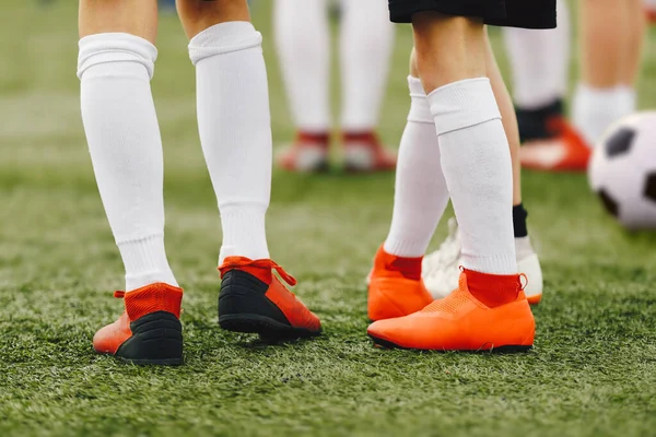 Legs of soccer players in red cleats on an artificial grass field. Football kids in a group on training. Sports white socks. Classic soccer ball in a background