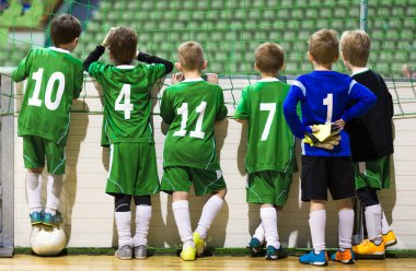 Kapalı okul futbol takımı. Çocuklar için futbol maçı. Arka planda spor salonu var. Çocuklar takım arkadaşlarını destekliyor. Çocuklar için kapalı futbol okulu turnuvası