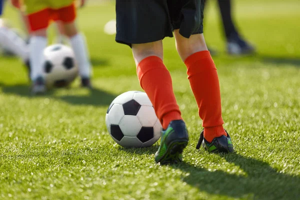 Outdoor Soccer Training For Kids. Legs of Kids Football Player Kicking Ball on Grass Venue. Children Soccer Summer School. Youth Athletes in Colourful Sports  Uniiforms and Soccer Cleats