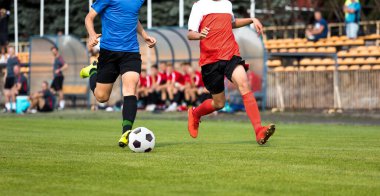 İki futbolcu düello yapıyor. Futbolcu gençler topta hızlı koşuyor. Oyuncular arka plandaki yedek bankta oturuyorlar. Kırmızı ve mavi formalı futbolcular