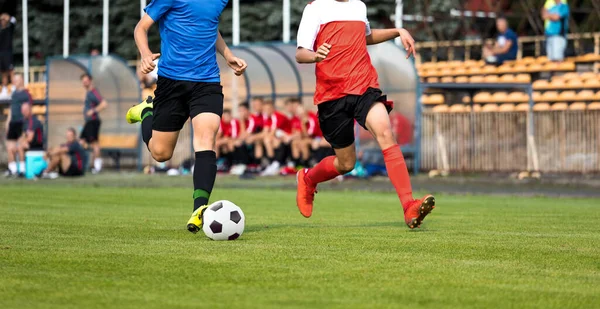 決闘を実行している2人のサッカー選手 サッカーの10代の少年たちはボールで速く走る 背景にはベンチに腰掛けてプレーヤー 赤と青のジャージのシャツを着たサッカー選手 — ストック写真