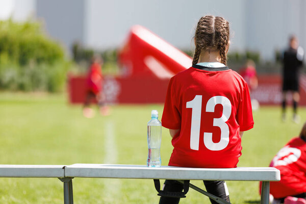 Girl Wearing Number 13 Jersey Sitting on Bench. Girl Soccer Player Resting on Sideline Bench. Youth Football Team Break with Number 13 Jersey and Water Bottle