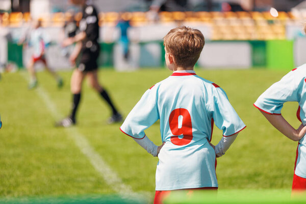 Boy in Light Blue Jersey Watching Soccer Match. Young Soccer Player Watching the Game. Boy in Light Blue Jersey Observes Football Match from Sidelines on Sunny Day