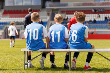 Mavi formalı Gençlik Futbol Takımı Stadyumdaki futbol maçı sırasında Sideline Bench 'te dinleniyor. Çocuklar Güneşli Gün 'de Futbol Ligi Oynuyor