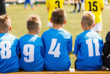 Maç sırasında ya da antrenman molasında mavi formalı çocuklar ve yedek kulübesinde oturan gençler.