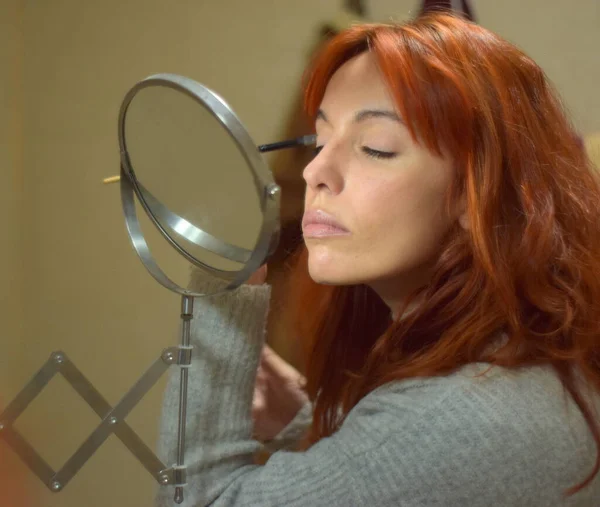 Young white woman with red hair puts on makeup in front of the mirror with the brush on her eyelashes