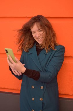 Genç kadın yeşil kaplamalı telefonunda gördüklerine şaşırmış bir şekilde gülümsüyor. Turuncu duvar arka planında turuncu bir arkaplan giyiyor ve yeşil kaplamalı telefonunda gördüklerine kibarca gülümsüyor. Turuncu duvar arkaplanı ve yeşil bir palto giyiyor.