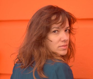 Kafkasyalı kadın saçları dalgalanırken kameraya derin derin bakar. Sokakta turuncu duvar arkaplanı