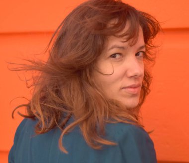 Kafkasyalı kadın saçları dalgalanırken kameraya derin derin bakar. Sokakta turuncu duvar arkaplanı