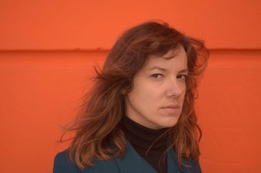 Beyaz kadın kameraya derinden bakıyor. Sokakta turuncu duvar arkaplanı