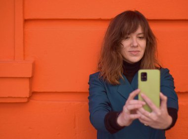 Arka planda turuncu duvar olan bir kadın selfie çekiyor. Mavi bir ceket giyiyor..