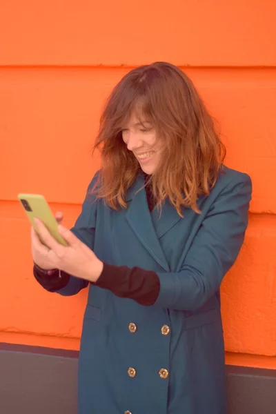 Genç bir kadın yeşil kaplamalı telefonunda gördüklerine nazikçe gülümsüyor. Turuncu duvar arkaplanı ve üzerinde deniz mavisi bir ceket var.