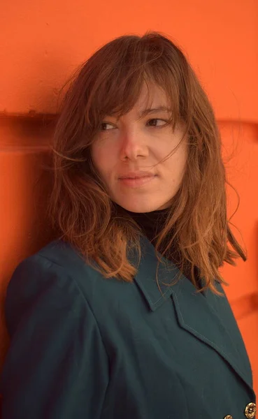 Turuncu bir duvara yaslanmış bir kadın ufka bakıyor. Mavi bir ceket giyiyor.