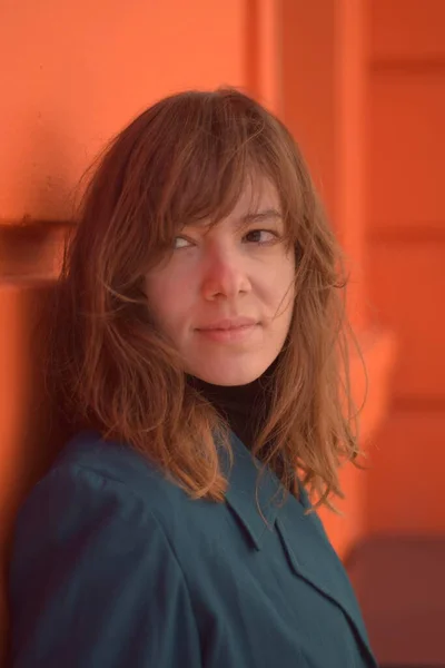 Turuncu bir duvara yaslanmış bir kadın ufka bakıyor. Mavi bir ceket giyiyor.