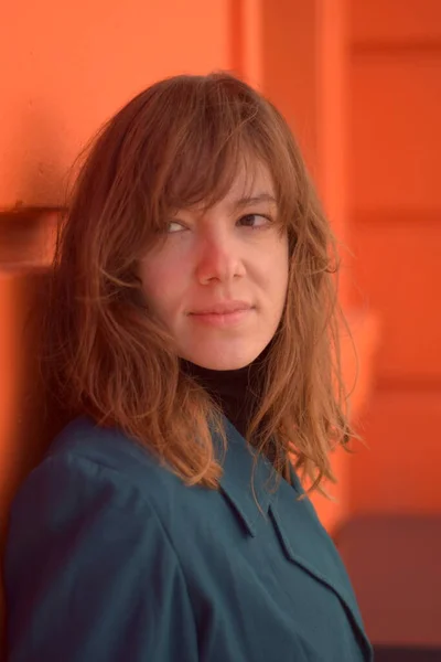 Turuncu bir duvara yaslanmış bir kadın ufka bakıyor. Mavi bir ceket giyiyor.