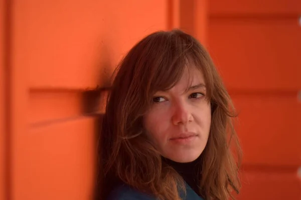 Turuncu bir duvara yaslanmış bir kadın ufka bakıyor. Mavi bir ceket giyiyor.