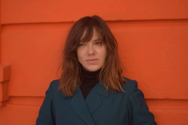 Önden çekilen mavi ceketli genç kadın turuncu arka planda gülümsüyor.