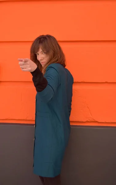 Kadın eliyle ateş etme hareketi yapıyor. Naranaj geçmişi olan kadın mavi bir ceket giyiyor..