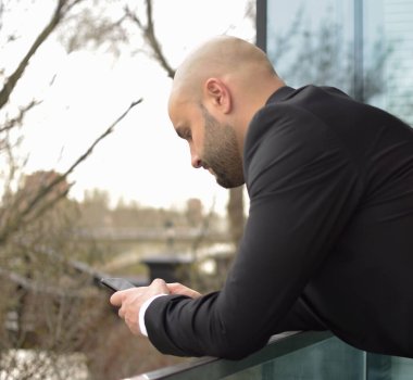 Takım elbiseli beyaz adam çite yaslanmış cep telefonuna bakıyor..