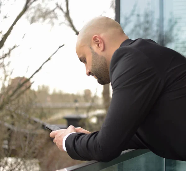 Takım elbiseli beyaz adam çite yaslanmış cep telefonuna bakıyor..