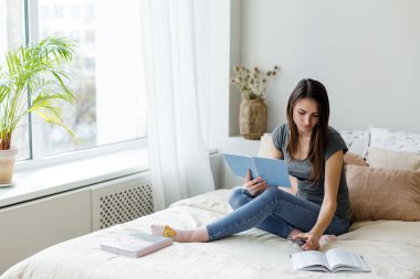 Yatakta oturup kitap okuyan ve telefona bakan bir kız..