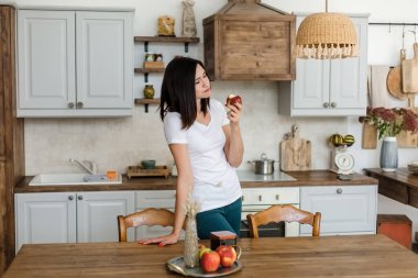 Mutfakta beyaz tişörtlü esmer kız elma yiyor..