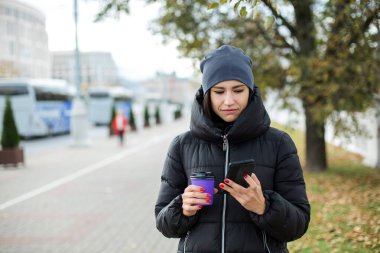 Ceketli ve şapkalı bir kız, elinde bir bardak ve bir telefon tutuyordu..
