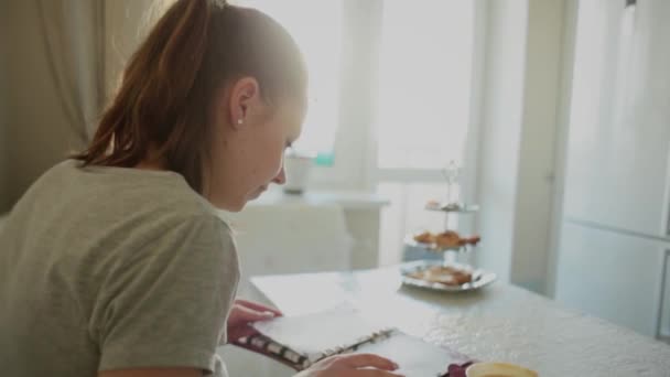 Κορίτσι κουζίνα απέναντι από το παράθυρο, στο τραπέζι, φύλλα μέσα από ένα σημειωματάριο — Αρχείο Βίντεο