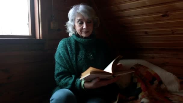 Äldre kvinna sitter i ett trärum och smickrar en bok. — Stockvideo