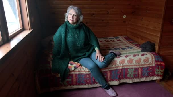 Mujer mayor se sienta en una habitación de madera y mira por la ventana sola. — Vídeo de stock