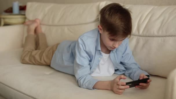 Niño se acuesta en el sofá en sus manos sosteniendo un teléfono, juega con él. — Vídeos de Stock