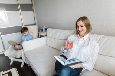 Kanepede oturan kadın elinde bir kitap tutuyor ve çay içiyor..