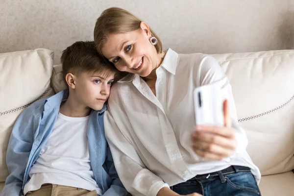 Chłopiec i dorosła kobieta robią sobie selfie przez telefon siedząc na kanapie. — Zdjęcie stockowe