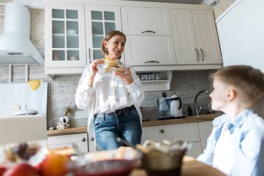 Yetişkin bir kadın mutfakta durur ve bir çocuğa krep ikram eder..