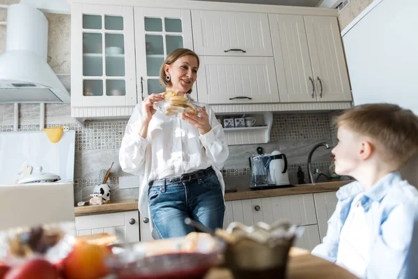 Yetişkin bir kadın mutfakta durur ve bir çocuğa krep ikram eder..