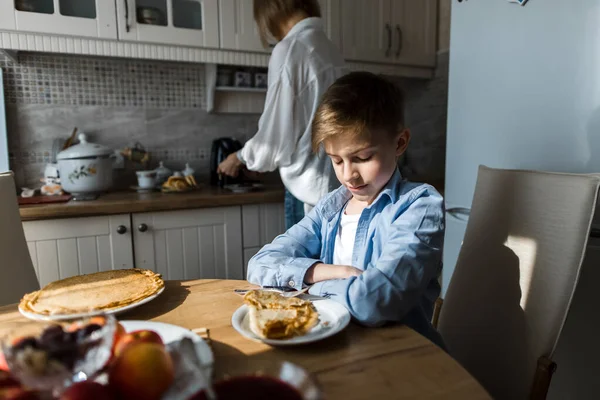 Gençler mutfaktaki masada oturur, arka plandaki kadın yemekleri hazırlar.