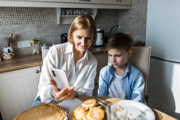 Nő a konyhában az asztalnál a gyermekkel nézi a telefont, reggeli az asztalon — Stock Fotó
