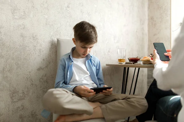 Çocuk mutfakta masada oturuyor ve telefonuna bakıyor..