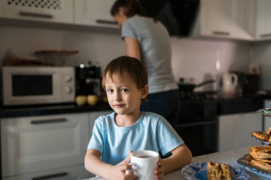 Çocuk elinde bir fincanla mutfak masasında oturur ve kameraya bakar..