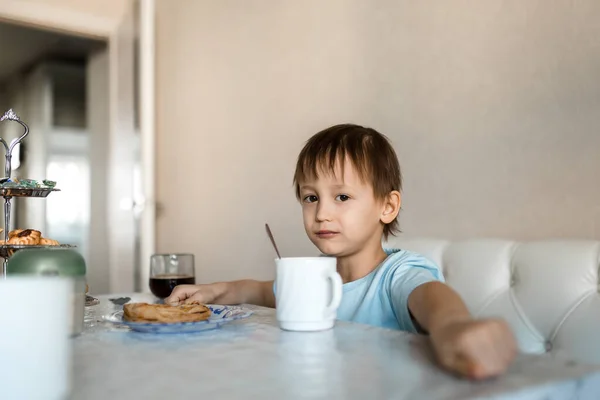 Çocuk mutfak masasında oturur ve kameraya, fincana ve tatlıya bakar.