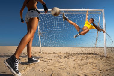 Kız topa vuruyor ve plajda kaleye asılıyor.