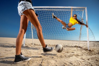 Kız topa vuruyor ve plajda kaleye asılıyor.