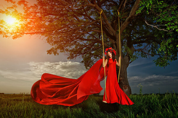 Beautiful woman in red dress with flower wreath on the swing in the forest in sunset