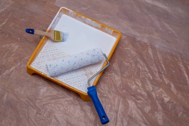 Koruma örtüsüyle kaplı bir zemindeki boya malzemeleri: beyaz boyalı bir boya tepsisi, bir boya silindiri ve bir boya fırçası.