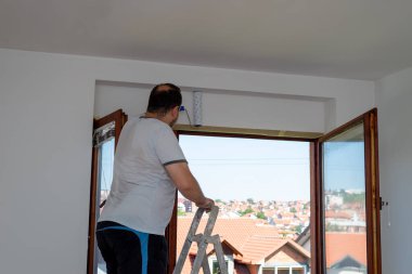 Merdivendeki adam pencerenin üstündeki duvarı beyaz renkte silindirle boyuyor. Pencere açık ve kırmızı çatılar görülebiliyor..